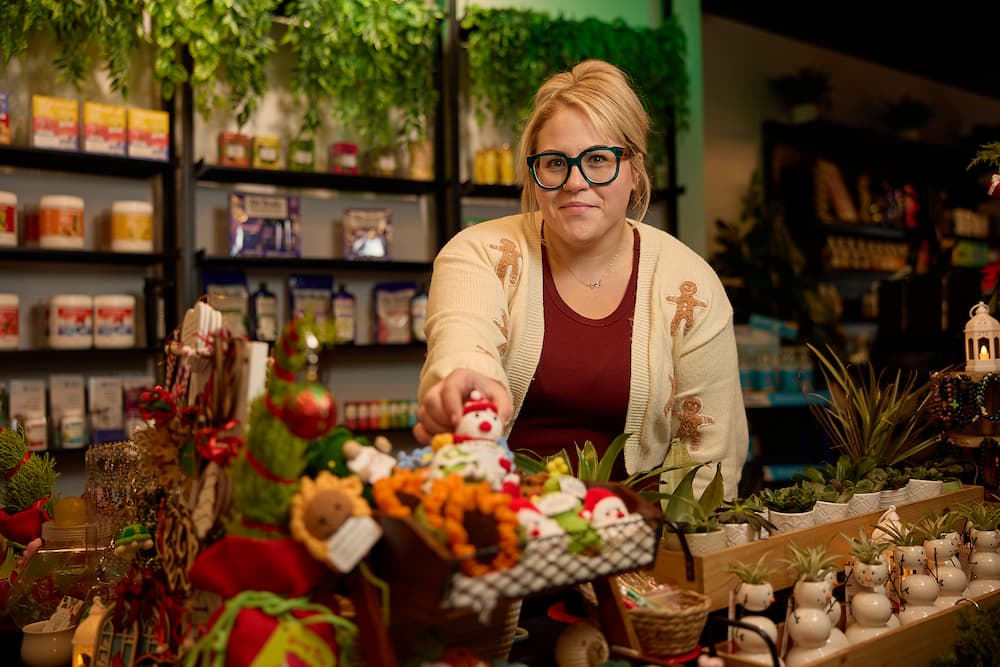 Andrea from Peach & Bloom, holding a small basket of colorful decorative items. Shelves with jars and plants are visible in the background.