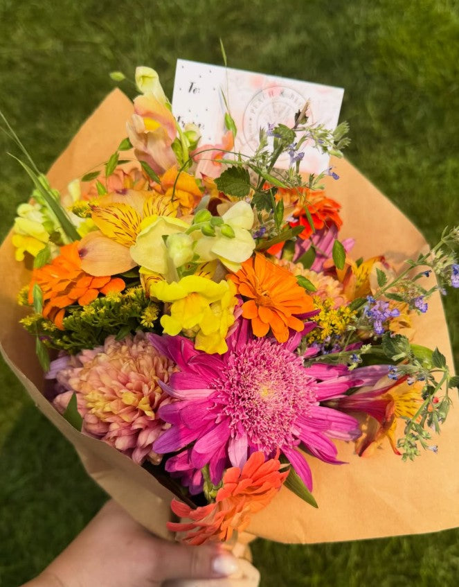 A vibrant, multicolor bouquet of fresh flowers wrapped in brown kraft paper, held by a hand against a green grass background. The bouquet features a mix of bright orange, yellow, pink, and purple flowers with green foliage and a small postcard.