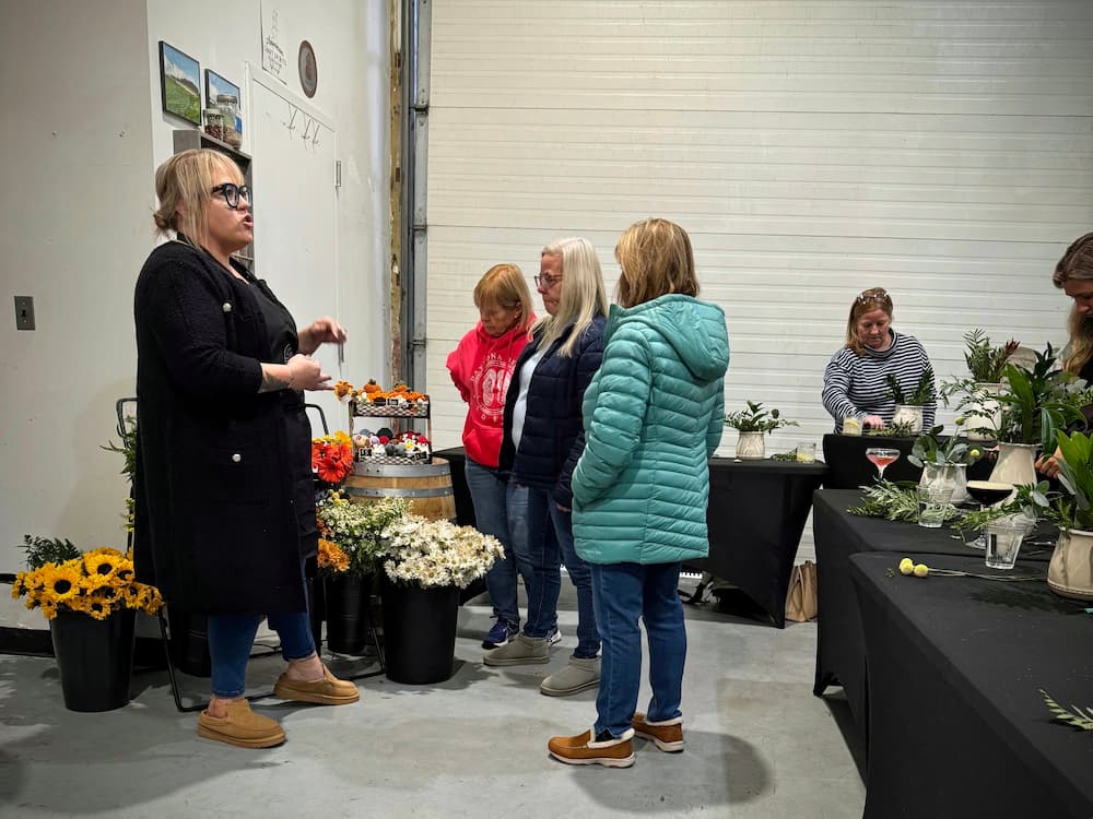 A woman in a black coat and glasses stands next to a flower arrangement display, talking to a group of people in an indoor event space with white garage door panels in the background. Workshops at Peach & Bloom in LI New York