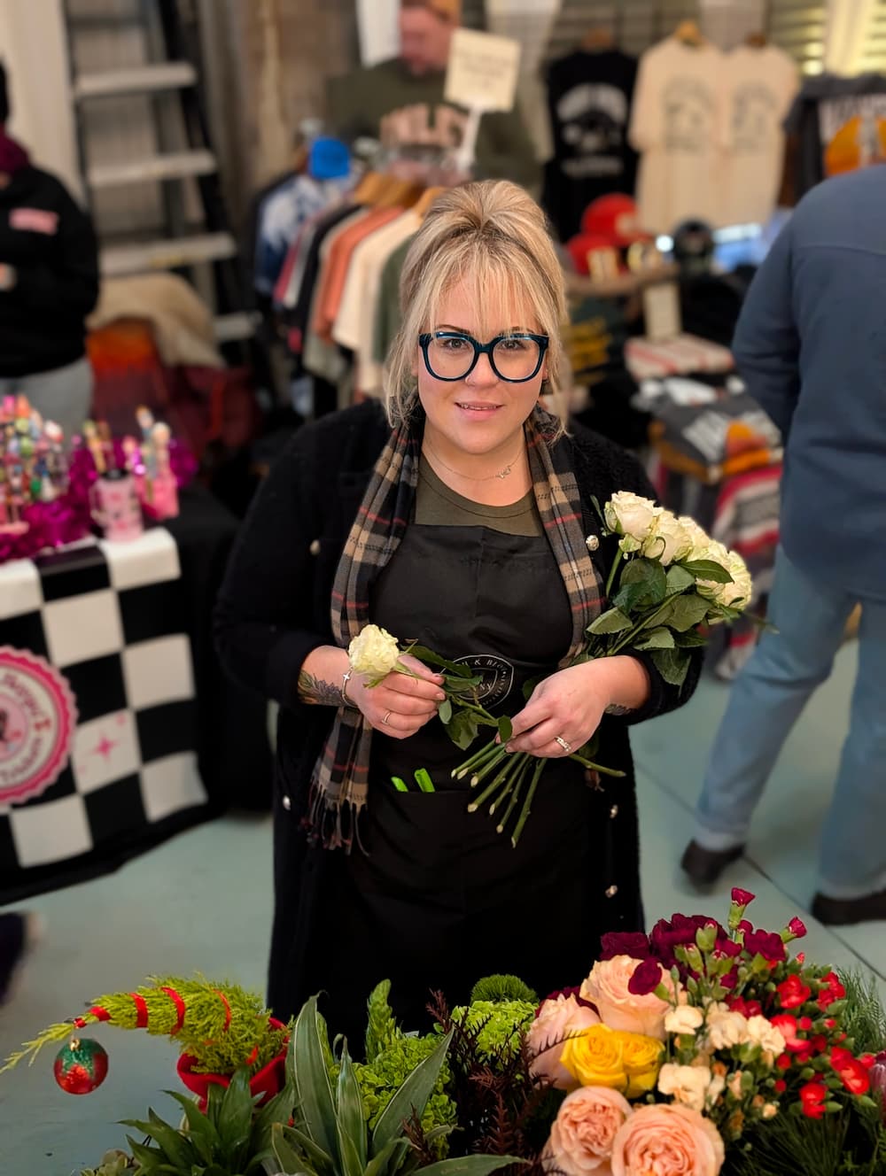 Andrea from Peach & Bloom holds a bouquet of white roses in a busy indoor market or shop setting with clothing racks and tables in the background. Long Island Event Florist in NY