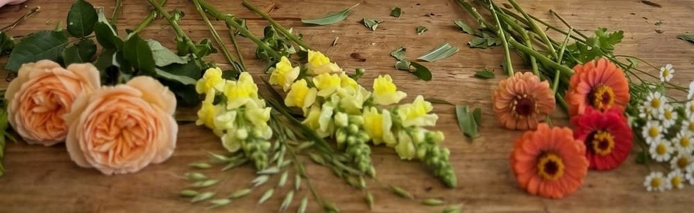 A flat-lay arrangement of fresh cut flowers on a rustic wooden surface, featuring peach roses, yellow snapdragons, orange and red zinnias, and small white daisies.