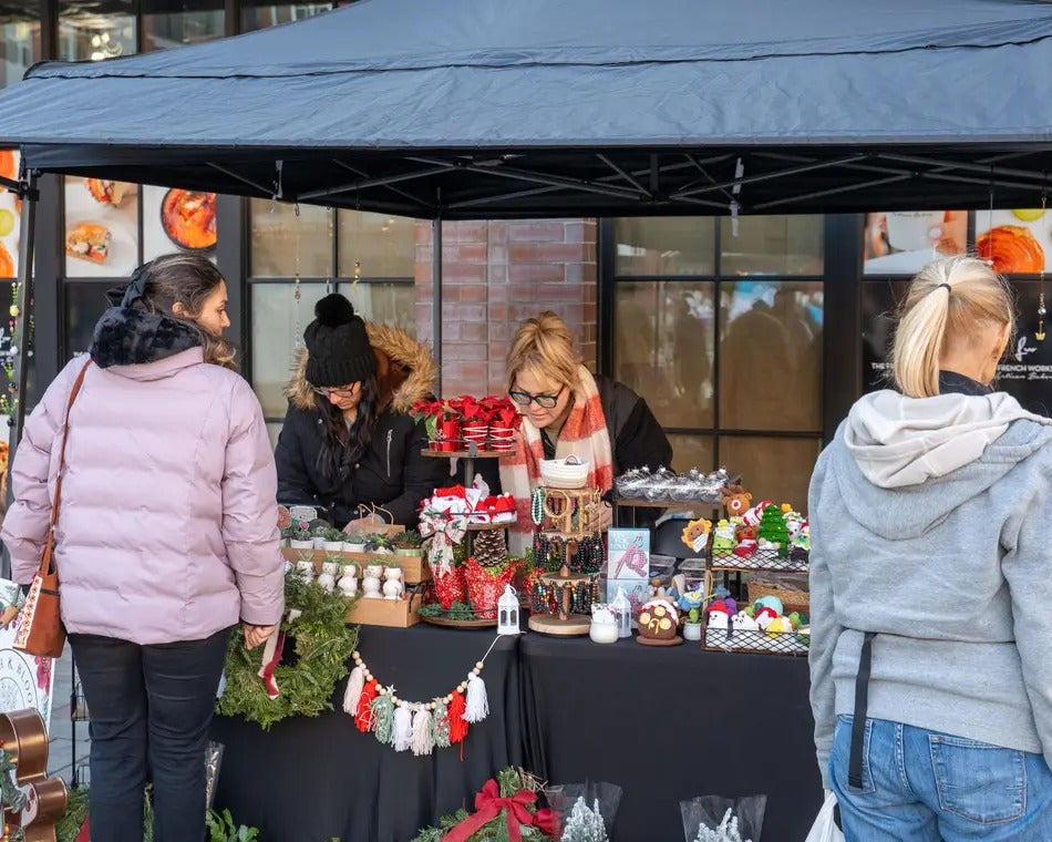 A market stall under a dark canopy with shoppers browsing handmade holiday decorations, ornaments, and small gift items. Several people are visible from the back and side, standing around the table with the stall staff behind it.