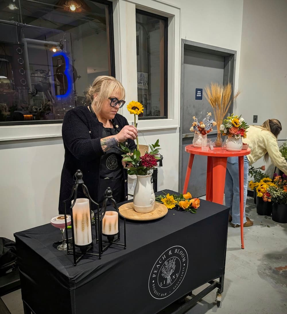 A woman arranging flowers at a craft booth table covered with a black cloth branded 'Black & White Floral Design', while another person works in the background near flower pots and a small red table with floral displays. Andrea from Peach & Bloom teaching a floral arranging workshop.